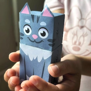 Op de afbeelding: Een blauw en grijs papieren kattenknutselwerk met een lachend gezicht, roze oren en een witte slabbetje. De kat wordt in iemands handen gehouden.