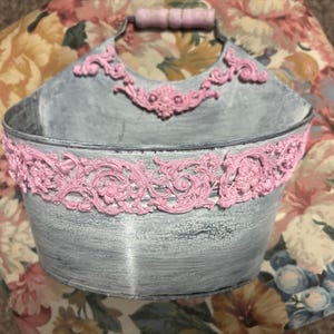 May include: A gray metal wall basket featuring a distressed finish and pink decorative scrollwork accents along the rim and handle. The basket has a semi-circular shape with a single handle at the top and a flat back for mounting purposes.