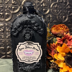 May include: A matte black decorative bottle featuring ornate raised scrollwork and a central label that reads Love Bites Brew, featuring a graphic of pink lips with fangs, the text Use With Caution, Lasts 12 Hours, A Sucker For Love, Love At First Fright.