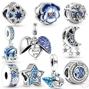 Op de afbeelding: Een verzameling zilveren bedels met blauwe en heldere kristallen accenten. Ontwerpen omvatten een bloem, een ster, een maan met sterren, een octopus, een zeeschelp en een hart met de tekst "I love you".