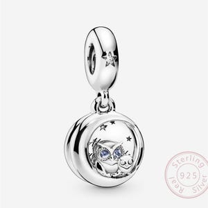 Könnte beinhalten: Ein silberner Charm mit einem Nachthimmel-Thema. Der Charm hat einen runden Anhänger mit einer Eule und Sternen in einem Halbmond. Ein kleiner Stern befindet sich auf der oberen Schlaufe. Der Charm ist aus Sterlingsilber gefertigt.