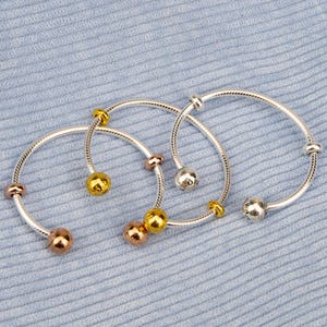 Puede incluir: Cuatro pulseras plateadas con cuentas doradas, oro rosa y plateadas. Las pulseras están sobre una superficie texturizada azul claro. Cada pulsera tiene un diseño de cuentas diferente, incluyendo formas redondas y cilíndricas.