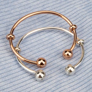 Könnte beinhalten: Zwei Armreifen, einer in Roségold und einer in Silber, werden auf einer hellblauen, strukturierten Oberfläche präsentiert. Der Roségold-Armreif hat eine glatte, polierte Oberfläche mit kugelförmigen Akzenten. Der Silberarmreif hat ein ähnliches Design mit silbernen Kugelakzenten.