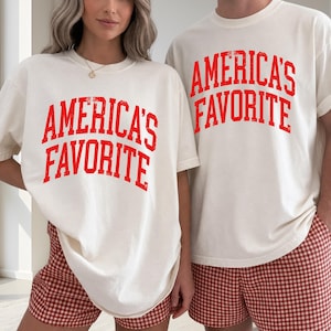 May include: Two off-white short-sleeve t-shirts featuring the red text "AMERICA'S FAVORITE" in a distressed, arched collegiate font. The shirts are paired with red and white gingham patterned shorts, creating a matching set of casual apparel.