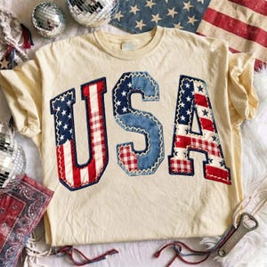 May include: A cream-coloured short-sleeve t-shirt featuring the letters USA appliqued on the front. The letters are constructed from a patchwork of blue fabric with white stars, red and white stripes, and red and white gingham check fabric.