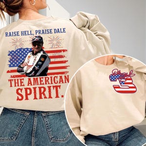 May include: Beige sweatshirt with the text "RAISE HELL, PRAISE DALE" and "THE AMERICAN SPIRIT." The design includes an American flag and a portrait of a man. A smaller design on the front features the number 3 and the word "Proud."