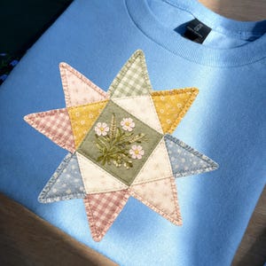 Peut inclure: Un sweat-shirt bleu clair à col rond orné d'une grande étoile en appliqué sur le devant. L'étoile est composée de divers empiècements en tissu aux tons vert sauge, jaune moutarde, rose poudré et bleu, avec des motifs floraux et vichy. L'empiècement central vert sauge est décoré de fleurs sauvages brodées.