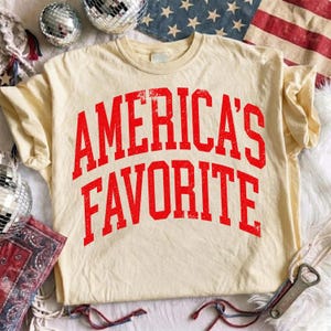 May include: A cream-colored short-sleeve t-shirt featuring the text AMERICA'S FAVORITE in a distressed, bold red collegiate-style font arched across the chest. The fabric has a soft, worn-in appearance with a classic crew neckline.
