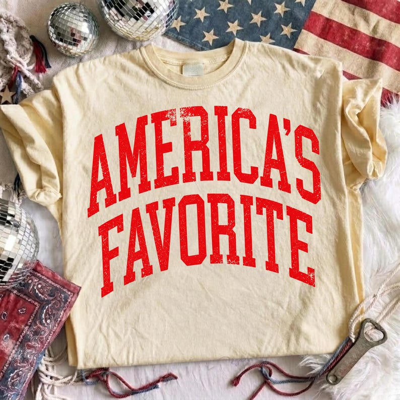 May include: A cream-colored short-sleeve t-shirt featuring the text AMERICA'S FAVORITE in a distressed, bold red collegiate-style font arched across the chest. The fabric has a soft, worn-in appearance with a classic crew neckline.
