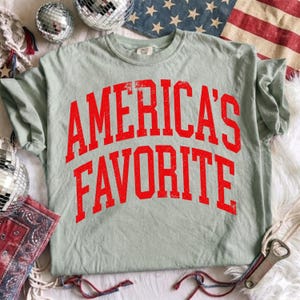 May include: A sage green short-sleeve t-shirt featuring the text "AMERICA'S FAVORITE" in a distressed red collegiate-style font across the chest. The shirt has a crew neckline and a relaxed fit, showcasing a textured, weathered appearance.