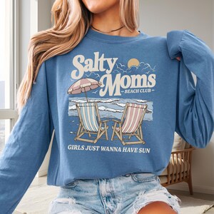 Op de afbeelding: Een blauw shirt met lange mouwen met een strandthema. De afbeelding bevat de tekst "Salty Moms Beach Club" boven een illustratie van twee strandstoelen, een parasol en oceaangolven. De tekst "GIRLS JUST WANNA HAVE SUN" staat eronder.