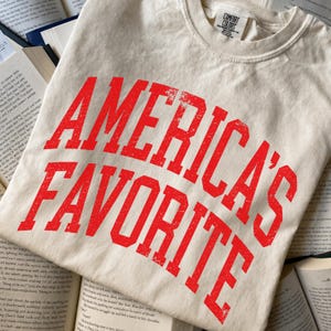 May include: A cream-colored short-sleeve t-shirt with a crew neckline. The front features the text "AMERICA'S FAVORITE" printed in a bold, distressed red font. The garment includes a Comfort Colors brand label inside the collar area.