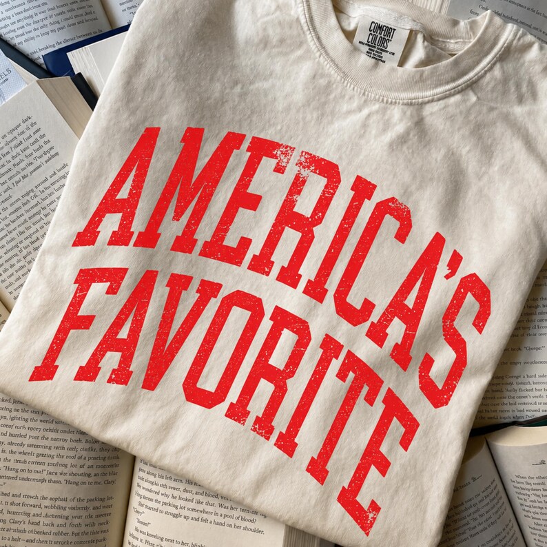 May include: A cream-colored short-sleeve t-shirt with a crew neckline. The front features the text "AMERICA'S FAVORITE" printed in a bold, distressed red font. The garment includes a Comfort Colors brand label inside the collar area.