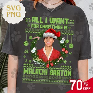 May include: A dark grey t-shirt with a Christmas design. The shirt displays the text "ALL I WANT FOR CHRISTMAS IS" in green, a portrait of a person in a Santa hat, and the name "MALACHI BARTON".