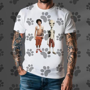 May include: White t-shirt featuring a graphic of two figures: a person in orange shorts and a towel, and an alien in red shorts. The shirt has a paw print pattern.
