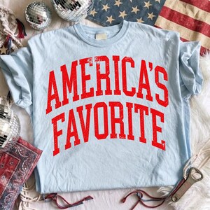 May include: A light blue short-sleeve t-shirt featuring the red text AMERICA'S FAVORITE in a distressed, arched varsity-style font across the chest. The shirt has a crew neckline and a standard fit, showcasing a casual and simple design.