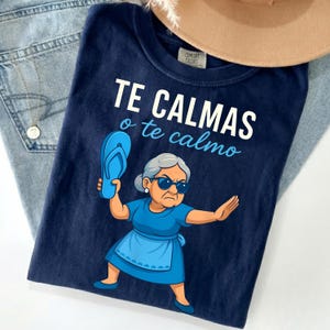 Könnte beinhalten: Marineblaues T-Shirt mit dem Text "TE CALMAS o te calmo" in Weiß und Hellblau. Die Grafik zeigt eine Cartoon-Oma, die eine blaue Flip-Flop hält. Ein beiger Hut ist im Hintergrund.