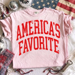 May include: A light pink short-sleeve t-shirt featuring the text AMERICA'S FAVORITE printed in a bold, distressed red font across the chest. The shirt has a crew neckline and a standard fit, showcasing a textured, weathered appearance on the lettering.