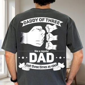 Può includere: T-shirt grigio scuro con grafica bianca. Il testo recita "Daddy of Three" sopra un pugno e tre pugni più piccoli. Sotto, c'è scritto "like a regular DAD, just three times as cool."