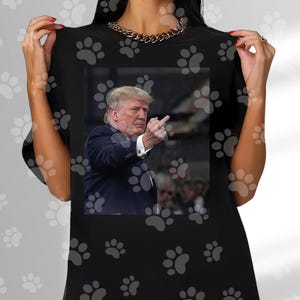 May include: Black t-shirt featuring a photographic image of a man in a suit, pointing his middle finger. The man has blonde hair and is in front of a blurred background. The shirt is plain black.