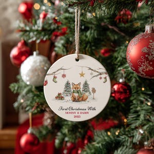 May include: Round Christmas ornament with a watercolor illustration of a fox wearing a green scarf, sitting in a snowy scene with Christmas trees and presents. The text reads "First Christmas With Mommy & Daddy 2023". The ornament hangs from a twine string.