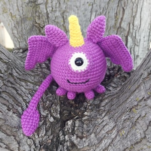 Puede incluir: Un monstruo de crochet morado con un cuerno amarillo, un ojo grande y una cola larga. El monstruo está sentado en un tronco de árbol.