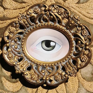 May include: A single eye painted in watercolor and framed in a gold ornate frame. The eye is green with a white highlight and a light brown iris.