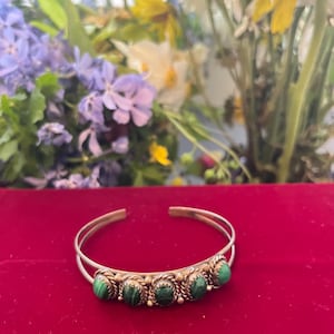 May include: Silver cuff bracelet featuring five green malachite stones. The bracelet has an open design with a row of round malachite stones set in a decorative metal band. The bracelet is displayed on a red surface.