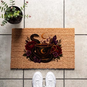May include: A rectangular tan coir doormat featuring a central graphic of a black teacup with a gold crescent moon, surrounded by a coiled black snake, deep red and purple flowers, purple crystal clusters, and a small gold crescent moon above.