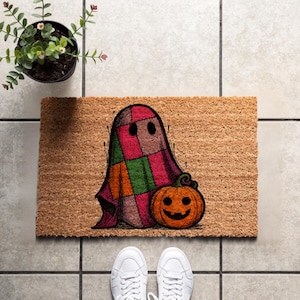 Peut inclure: Un paillasson rectangulaire en fibre de coco beige avec une illustration imprimée d'un fantôme recouvert d'un motif de courtepointe en patchwork multicolore. Le fantôme est placé à côté d'une citrouille orange avec un visage sculpté.