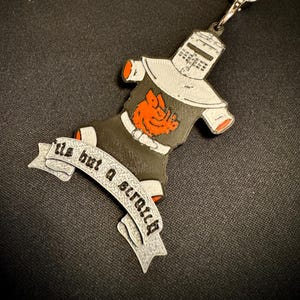 Può includere: Un portachiavi con un cavaliere in armatura nera e grigia con dettagli arancioni e un emblema rosso. Il portachiavi ha uno striscione che recita "tis but a scratch". Le parole "Monty Python" sono arcuate sopra il portachiavi.