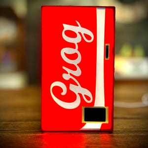 Könnte beinhalten: Eine rote, rechteckige Leuchtbox mit dem Wort "Crog" in weißer Schrift. Ein weißer vertikaler Streifen verläuft die rechte Seite hinunter. Ein kleines schwarzes Rechteck befindet sich unten, goldfarben umrahmt. Die Box steht auf einer Holzoberfläche.