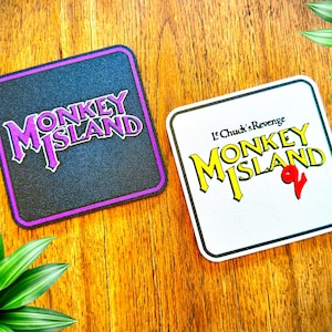 Könnte beinhalten: Zwei quadratische Untersetzer mit dem Text "Monkey Island" in einer stilisierten Schriftart. Ein Untersetzer ist schwarz mit einem lila Rand, der andere ist weiß mit gelbem Text und dem Text "LeChuck's Revenge". Die Untersetzer liegen auf einer Holzoberfläche.