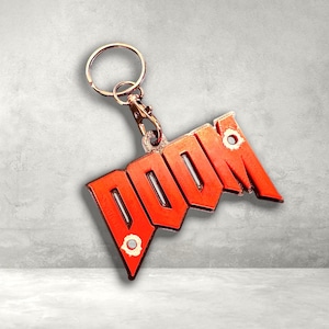 May include: A red and orange metal keychain featuring the word "DOOM" in a distinctive font. The keychain includes a metal ring and clasp. The backdrop is a light grey concrete wall.