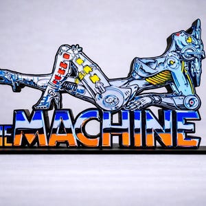 Peut inclure: Sculpture métallique stylisée d'une figure robotique allongée, avec des accents bleus, argentés, jaunes et rouges. La sculpture est montée sur une base noire avec l'inscription "THE MACHINE" en orange et bleu.
