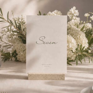 Pode incluir: Um cartão de número de mesa com a palavra "Seven" em escrita elegante. O cartão está sobre uma base de pedra, com flores brancas e vegetação ao fundo. O cartão também tem o texto "REBECCA & RIE" na parte inferior.