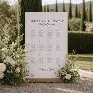Puede incluir: Un cartel rectangular blanco para el plan de asientos de una boda con texto en negro que dice OUR FAVORITE PEOPLE, Please find your seat, Rebecca & Steven, y 2.26.27 NEW YORK, NY, con columnas de nombres organizadas de la A a la Z sobre una base de madera.
