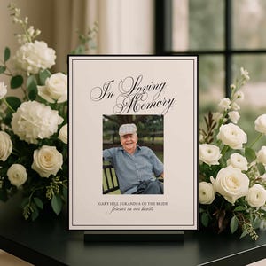 May include: A rectangular memorial sign with a cream background and black border. The text reads In Loving Memory, Gary Hill | Grandpa of the Bride, and forever in our hearts. A centered photograph shows an older person wearing a cap and shirt.