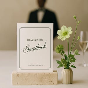 May include: A wedding guestbook sign on a beige stone block. The sign reads "PLEASE SIGN OUR Guestbook" in elegant script, with the names "HANNAH & SCOTT 06.25" below. A small vase with green flowers sits nearby.