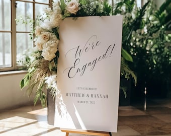 May include: A white sign on a wooden easel with the words "We're Engaged!" in elegant script. Below, it reads "LET'S CELEBRATE!" followed by names and a date. The sign is adorned with white and cream-colored flowers and greenery.