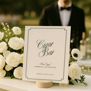 Könnte beinhalten: Ein weißes Schild mit der Aufschrift "Cigar Bar" in eleganter Schrift, umrahmt von einem grünen Rand. Das Schild steht auf einem kleinen, runden, beigen Ständer und ist von weißen Rosen umgeben. Der Text "Please Enjoy!" und Namen sind ebenfalls sichtbar.
