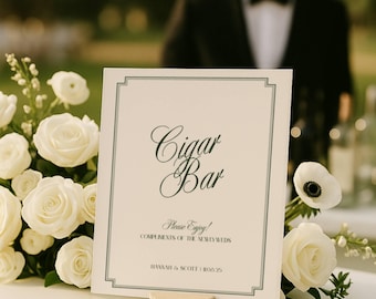 May include: A white sign with the words "Cigar Bar" in elegant script, framed with a green border. The sign is propped up on a small, round, beige stand and surrounded by white roses. The text "Please Enjoy!" and the names "Hannah & Scott" are also visible.