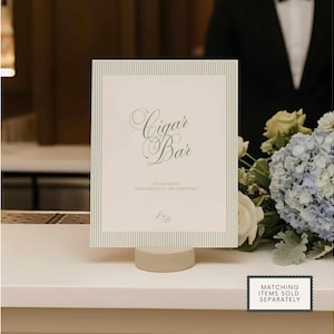 Puede incluir: Un letrero rectangular con las palabras "Cigar Bar" en escritura elegante, enmarcado por un borde a rayas. El letrero est&aacute; apoyado en un peque&ntilde;o soporte redondo beige. Un ramo de flores est&aacute; en el fondo.
