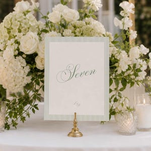 Puede incluir: Una tarjeta rectangular de número de mesa con la palabra Seven en letra cursiva verde, enmarcada por finas rayas verticales verdes sobre fondo blanco, sostenida por un pequeño soporte de metal dorado entre arreglos florales blancos.