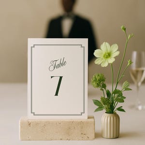 Puede incluir: Una tarjeta de mesa con el número 7, con un borde decorativo, exhibida sobre un bloque de piedra beige. Un pequeño jarrón con flores verdes y una copa de champán sugieren una boda o evento formal.