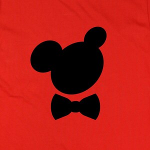 May include: A bright red t-shirt featuring a black silhouette of a cartoon character's head and bow tie. The design is centered on the shirt, offering a playful and recognizable graphic. The shirt is made of a soft material.