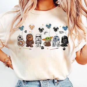 May include: Cream-coloured t-shirt featuring cartoon characters from Star Wars, each holding a balloon with a Mickey Mouse head design. Characters include BB-8, Chewbacca, a Stormtrooper, Grogu, R2-D2, and Darth Vader. The shirt has a relaxed fit.