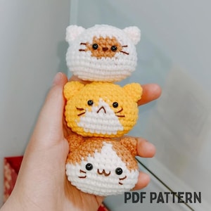 May include: Three crocheted cat-shaped ornaments stacked vertically. The top cat is white with brown markings, the middle is yellow with black details, and the bottom is white and brown. Each has black eyes and whiskers. The text "PDF PATTERN" is visible.