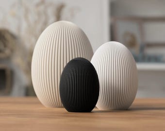 Può includere: Tre oggetti decorativi a forma di uovo con dettagli a coste verticali. Due sono bianco sporco e uno è nero. Sono disposti su una superficie di legno, creando un'estetica minimalista. Le uova variano in dimensioni.