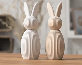 Può includere: Due statuine di coniglio in ceramica, una bianca e una beige, con una texture a coste. I conigli hanno orecchie lunghe e sono in piedi su una superficie di legno.